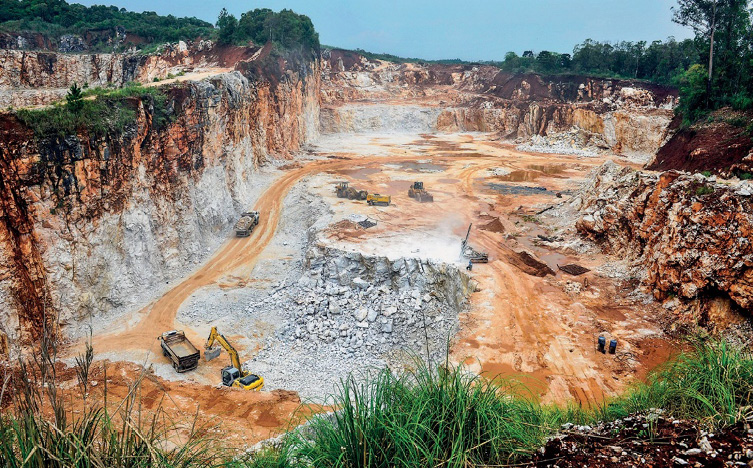 Imagem: Fotografia. Caminhões em um terreno com pedras. Em volta há paredes de terra. Fim da imagem.