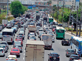 Imagem: Fotografia. Uma fileira de carros, caminhões e ônibus parados em uma avenida. Ao fundo, prédios e árvores. Fim da imagem.