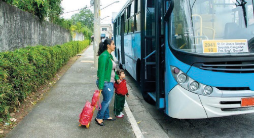 Imagem: Fotografia. Uma mulher está segurando a mão de um menino e com a outra mão, ela segura uma mochila de rodinhas. Na frente deles há um ônibus parado com a porta aberta.  Fim da imagem.
