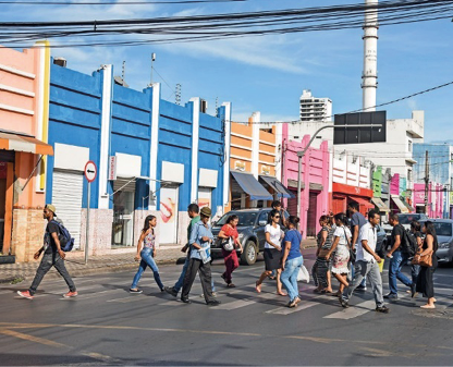 Imagem: Fotografia. Várias pessoas estão atravessando uma rua sobre a faixa de pedestres. Atrás delas há carros parados e ao fundo, construções coloridas. Fim da imagem.