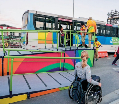 Imagem: Fotografia. Uma mulher em cadeira de rodas está descendo uma rampa. Ao fundo há um ônibus colorido parado com a porta aberta. Fim da imagem.