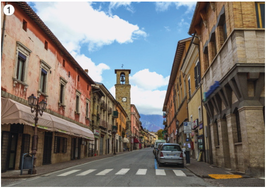 Imagem: Fotografia 1. No centro há carros em uma rua estreita. Nas laterais há construções extensas e altas.   Fim da imagem.