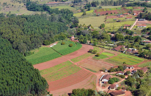 Imagem: Fotografia. Vista aérea de uma plantação extensa ao lado de casas espalhadas. Ao fundo, muitas árvores. Fim da imagem.