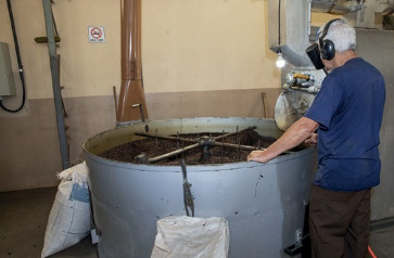 Imagem: Fotografia. Um homem com fone na cabeça está com as mãos sobre um barril grande e cinza. Ao lado há duas sacas encostadas. Fim da imagem.