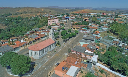 Imagem: Fotografia. Vista aérea de uma igreja e árvores. Em volta há uma rua e casas. Ao fundo, construções e mais árvores.  Fim da imagem.