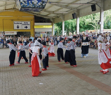 Imagem: Fotografia. Meninas com chapéu e camiseta branca e saia comprida e vermelha estão com as mãos nas cinturas. Entre elas, meninos com lenço vermelho em volta do pescoço, camisa xadrez, chapéu e calça preta estão de mãos dadas. Ao fundo, uma multidão observa. Fim da imagem.