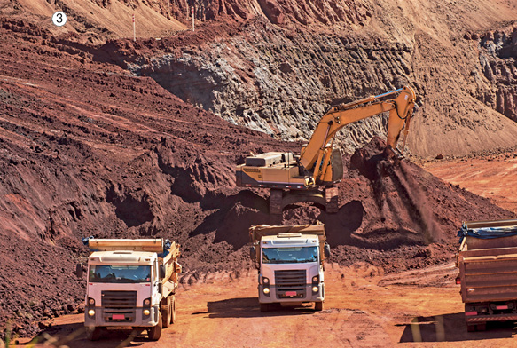 Imagem: Fotografia 3. Dois caminhões e uma escavadeira em um terreno com terra escura. Fim da imagem.