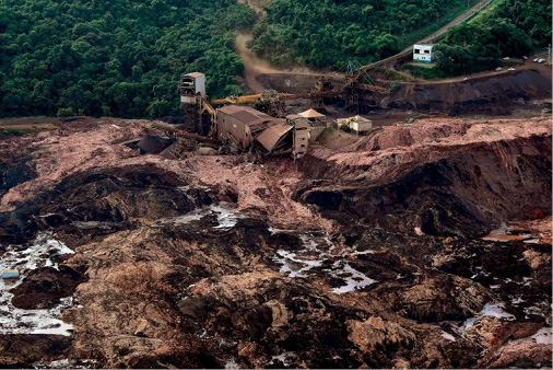 Imagem: Fotografia. Vista aérea de uma casa destruída com lama em volta. Ao fundo, plantas e árvores. Fim da imagem.