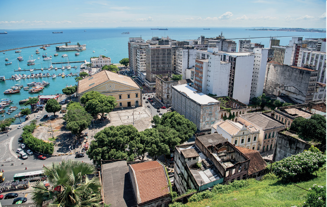 Imagem: Fotografia. No centro há várias árvores, casas e prédios. Ao fundo, barcos atracados em um porto no mar azul. Fim da imagem.