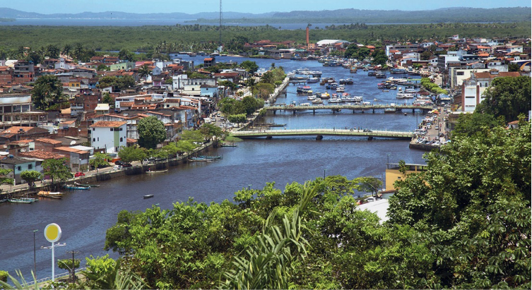Imagem: Fotografia. No centro há um rio extenso e largo com duas pontes e vários barcos. Nas margens há árvores e construções. Ao fundo, mais árvores e morros. Fim da imagem.