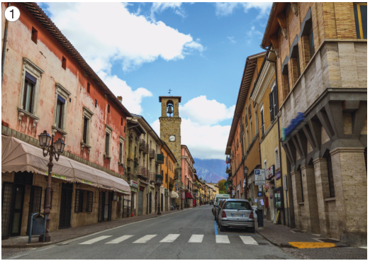Imagem: Fotografia 1. No centro há carros em uma rua estreita. Nas laterais há construções extensas e altas.   Fim da imagem.