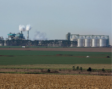 Imagem: Fotografia. No centro há um terreno plano e verde e ao fundo, uma usina grande com chaminés altas, expelindo fumaça branca.  Fim da imagem.