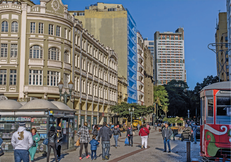 Imagem: Fotografia. No centro há pessoas andando na calçada. À esquerda, prédios e construções e à direita, um bonde na rua. Fim da imagem.