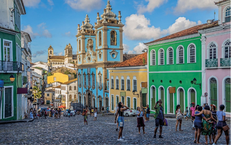 Imagem: Fotografia. Pessoas andando em uma rua de pedras. Em volta há construções com paredes coloridas. Fim da imagem.