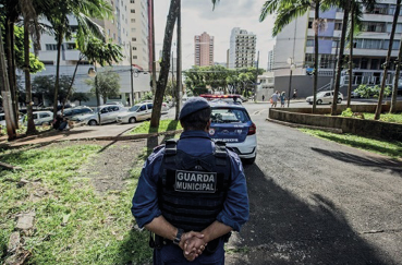 Imagem: Fotografia. Um homem com quepe e uniforme azul-escuro e colete preto com a informação: GUARDA MUNICIPAL está com as mãos atrás das costas. Na frente dele há um carro de polícia em uma praça. Ao fundo, carros, árvores e construções. Fim da imagem.