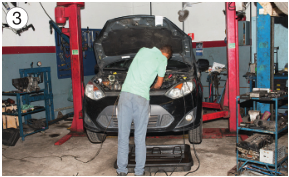 Imagem: Fotografia 3. Um homem está de costas e mexendo no capô de um carro preto. Em volta dele há várias ferramentas.   Fim da imagem.