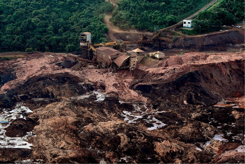 Imagem: Fotografia. Vista aérea de uma casa destruída com lama em volta. Ao fundo, plantas e árvores. Fim da imagem.