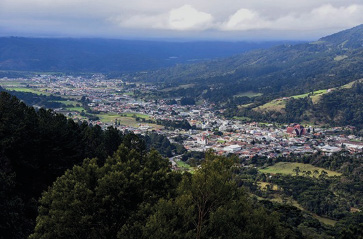 Imagem: Fotografia. Vista aérea de construções entre morros verdes. Fim da imagem.