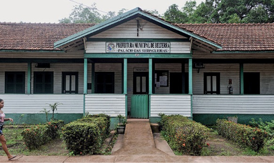 Imagem: Fotografia. Construção com parede branca e verde. Em volta há muitas plantas e ao fundo, árvores. Fim da imagem.