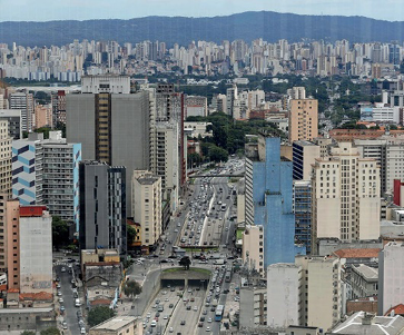 Imagem: Fotografia. Vista aérea de uma avenida com automóveis. Nas laterais há vários prédios.  Fim da imagem.