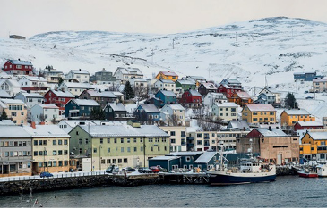 Imagem: Fotografia 2 (ZONA POLAR ÁRTICA). Barcos e embarcações em um porto. Ao lado, construções e ao fundo, morros brancos.  Fim da imagem.