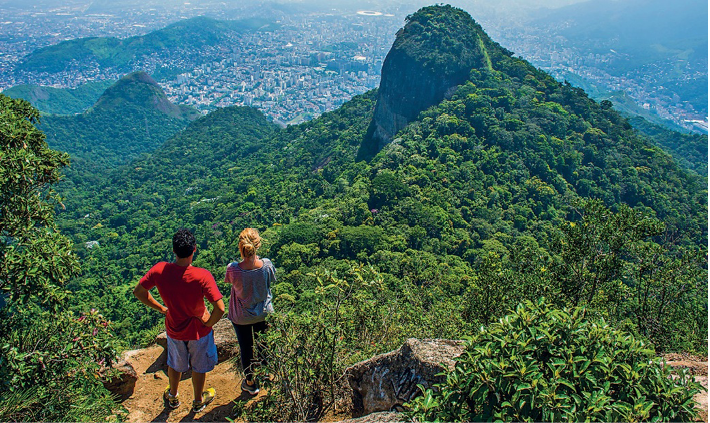 Imagem: Fotografia. Um casal está parado no topo de um morro. Abaixo deles há um morro coberto de árvores e ao fundo, uma cidade. Fim da imagem.