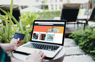 Imagem: Fotografia. Mão direita de uma pessoa sobre um teclado de notebook e a mão esquerda está segurando um cartão de crédito. Na frente da pessoa há um site de compras na tela do notebook.   Fim da imagem.