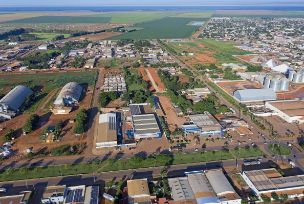 Imagem: Fotografia. À esquerda, galpões grandes entre árvores. No centro, construções e ruas e à direita, uma usina. Em volta há casas, ruas e ao fundo, terrenos verdes. Fim da imagem.