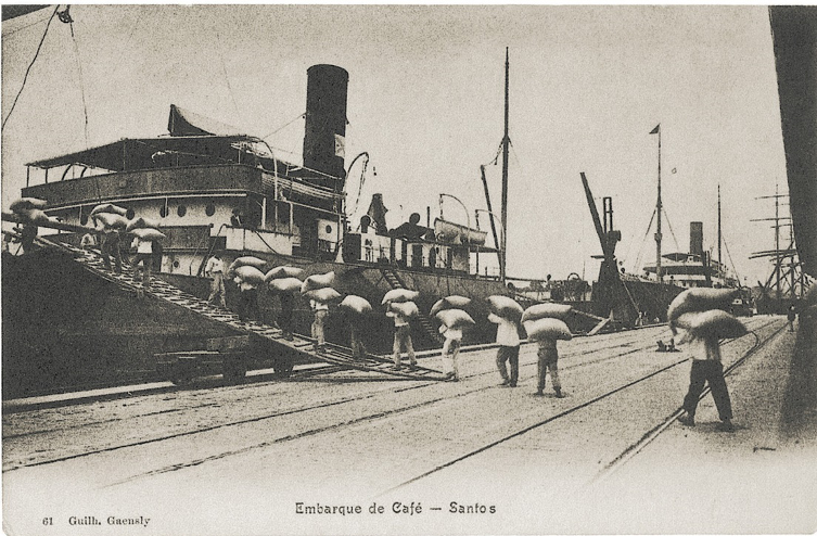 Imagem: Fotografia em preto e branco. Homens estão enfileirados, segurando sacas grandes sobre as cabeças e subindo em um navio. Ao fundo, mais navios atracados. Fim da imagem.