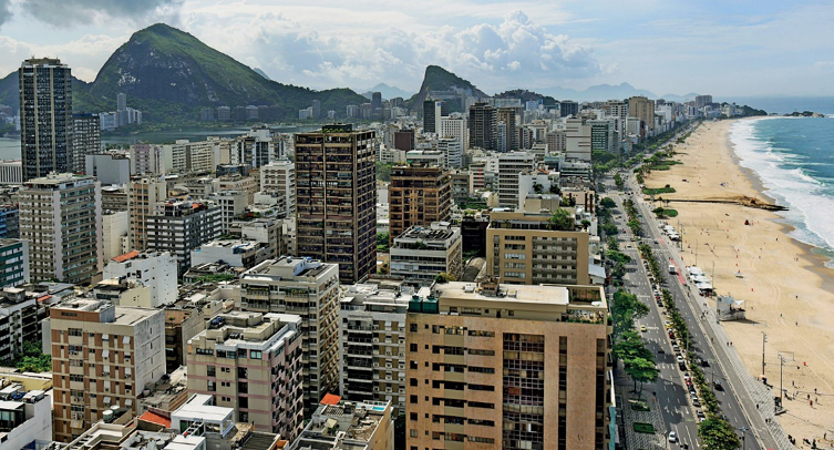 Imagem: Fotografia. Vista aérea da orla da praia. À esquerda, uma rua asfaltada e prédios. Ao fundo, morros. Fim da imagem.