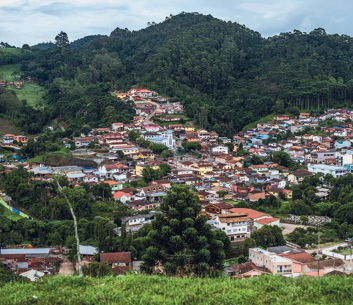 Imagem: Fotografia. Vista aérea de uma cidade entre morros verdes.  Fim da imagem.