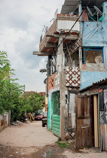 Imagem: Fotografia. Casa improvisada com três andares. Ao fundo, mais casas, árvores e carros.  Fim da imagem.