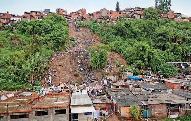 Imagem: Fotografia. Casas embaixo de um morro. Acima há um deslizamento e árvores. Na parte superior do morro há mais casas.  Fim da imagem.