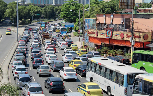 Imagem: Fotografia. Vários veículos enfileirados em uma avenida. Nas laterais há árvores e construções.  Fim da imagem.