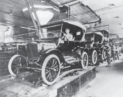 Imagem: Fotografia em preto e branco. Uma fileira de carros sobre uma esteira. Ao lado há homens.   Fim da imagem.