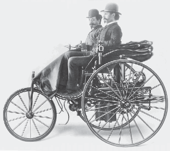 Imagem: Fotografia em preto e branco. Dois homens com chapéu e terno estão sentados em um veículo com três rodas, duas grandes na parte de trás e uma média na frente.   Fim da imagem.