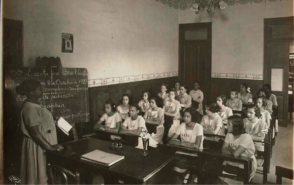 Imagem: Fotografia em preto e branco. Uma professora está em pé e segurando um livro. Na frente dela, várias meninas estão sentadas em duplas, em carteiras escolares e observando.  Fim da imagem.
