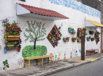 Imagem: Fotografia. Parede branca com desenhos de árvores e flores coloridas. Nela há vasos pendurados com plantas. Ao lado, um banquinho amarelo e um ponto de ônibus. Fim da imagem.