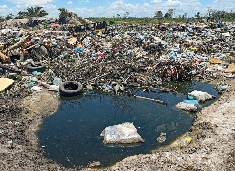 Imagem: Fotografia. Muito lixo em volta de uma poça d’água escura. Fim da imagem.