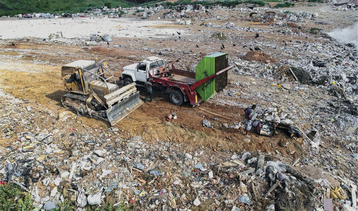 Imagem: Fotografia. Um caminhão está despejando lixo sobre um terreno e ao lado, um trator está cobrindo o terreno com terra. Em volta há muito lixo espalhado.  Fim da imagem.