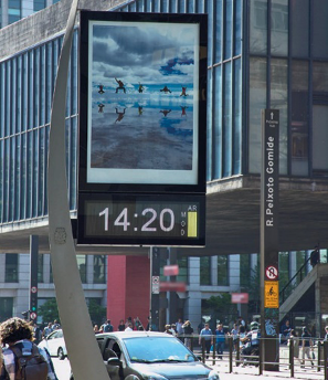 Imagem: Fotografia. Um painel com uma foto e abaixo há um relógio indicando 14:20 horas e ao lado, a informação: AR – MOD e um retângulo amarelo. Ao fundo, carros, pessoas e construções.  Fim da imagem.