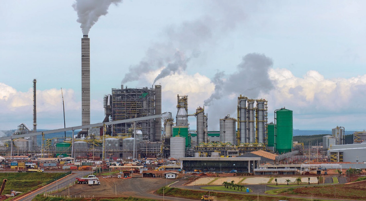 Imagem: Fotografia. Uma indústria com chaminés expelindo fumaça preta.  Fim da imagem.