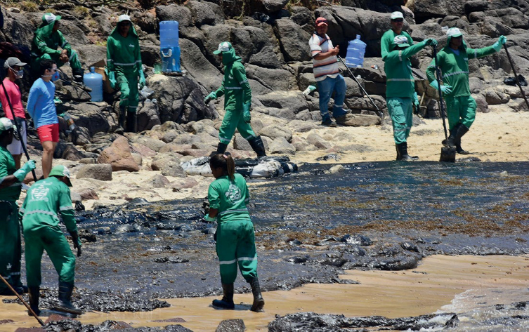 Imagem: Fotografia. Várias pessoas com roupas verdes e botas estão segurando vassouras. Entre elas há uma mancha preta na areia da praia. Ao fundo, rochas e mais pessoas.   Fim da imagem.