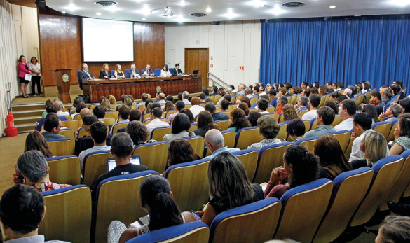 Imagem: Fotografia. Várias pessoas estão sentadas em um auditório. Ao fundo, sete pessoas estão sentadas atrás de uma mesa comprida. Atrás delas há uma tela de projeção.  Fim da imagem.