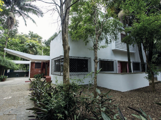 Imagem: Fotografia. Construção com paredes brancas e em volta há várias árvores e plantas.  Fim da imagem.