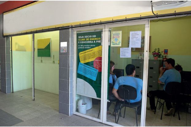Imagem: Fotografia. Salas com pessoas no interior e porta de vidro com visão par ao interior da sala. No interior há uma mulher e dois homens sentados, vestindo camiseta azul. Fim da imagem.