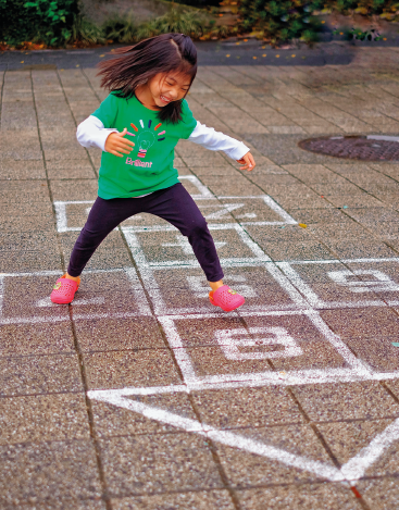 Imagem: Fotografia. Menina com cabelos escuros e lisos na altura dos ombros, sorrindo e com os olhos fechados. Veste camiseta verde, calça roxa e chinelo rosa. Está saltando com um pé na direção de quadrados rabiscados no chão com tinta branca. Dentro dos quadrados há números e um triângulo abaixo do quadrado com o número 8. Ao fundo, grama curta e concreto.  Fim da imagem.