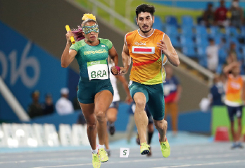 Imagem: Fotografia. À esquerda, uma mulher, com uma venda verde e amarela nos olhos, camisa em tons de verde com placa de identificação escrito BRA, corre e segura, com a mão direita, um bastão amarelo. À sua direita, um homem segura a mão esquerda dela enquanto corre ao seu lado. Ao fundo, pessoas e uma arquibancada.  Fim da imagem.