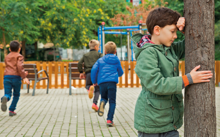 Imagem: Fotografia. Uma criança, com cabelo curto e blusa verde, de olhos fechados, apoia o rosto no tronco de uma árvore. Ao fundo, outras crianças correm. Em seguida, há cadeiras, cercas e árvores.  Fim da imagem.