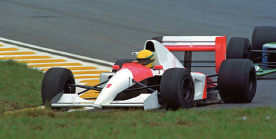 Imagem: Fotografia. Um carro de automobilismo, branco e vermelho. Dentro está o motorista com um capacete amarelo, está na grama. Ao fundo, uma pista cinza.   Fim da imagem.